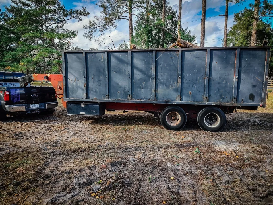High sided dump trailer hitched to a pickup on a cleared lot with a log in the load, debris removal