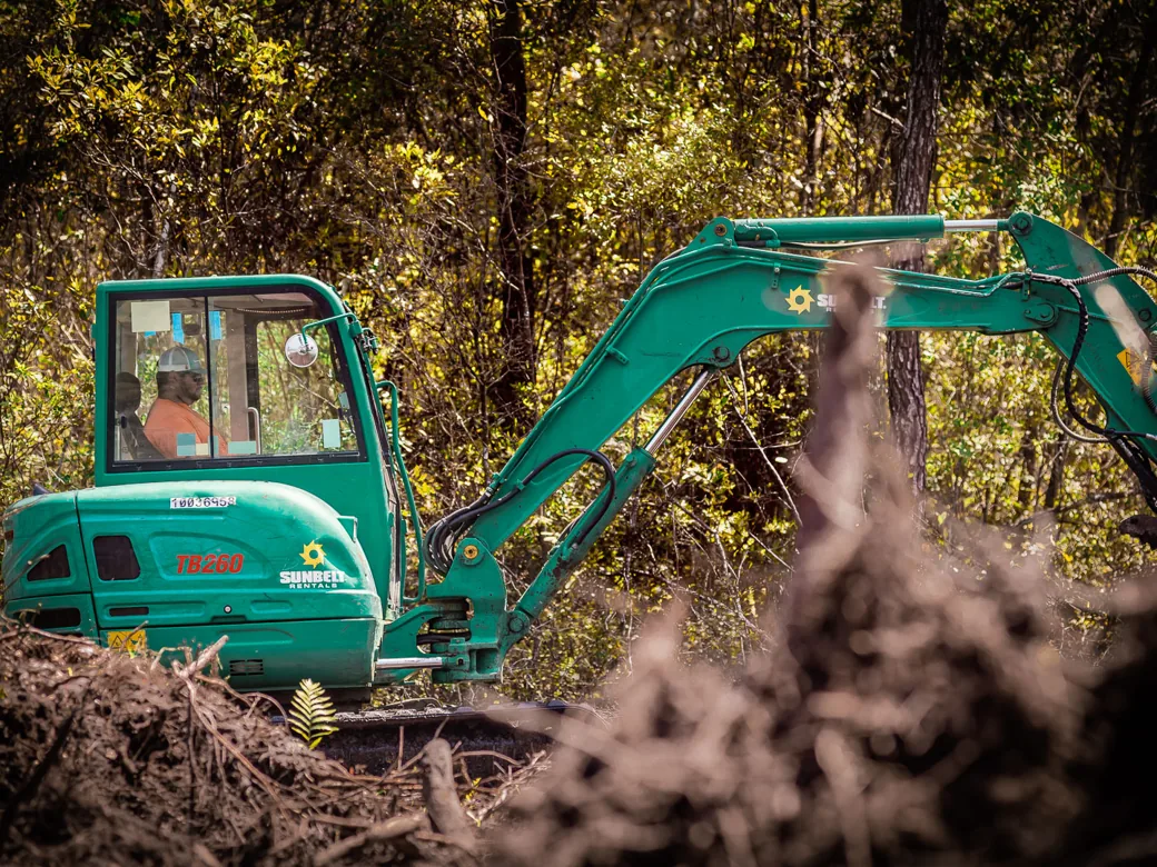 Excavator working beyond piles of pulled roots and brush, debris removal