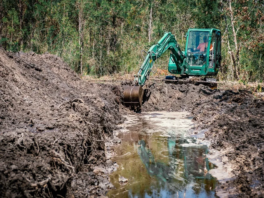 Excavator digging a long pond with water filling the cut and spoil banked on both sides, excavating