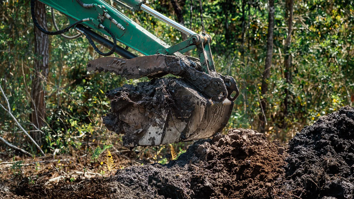 Excavator bucket full of muck over a spoil pile in the woods, land clearing