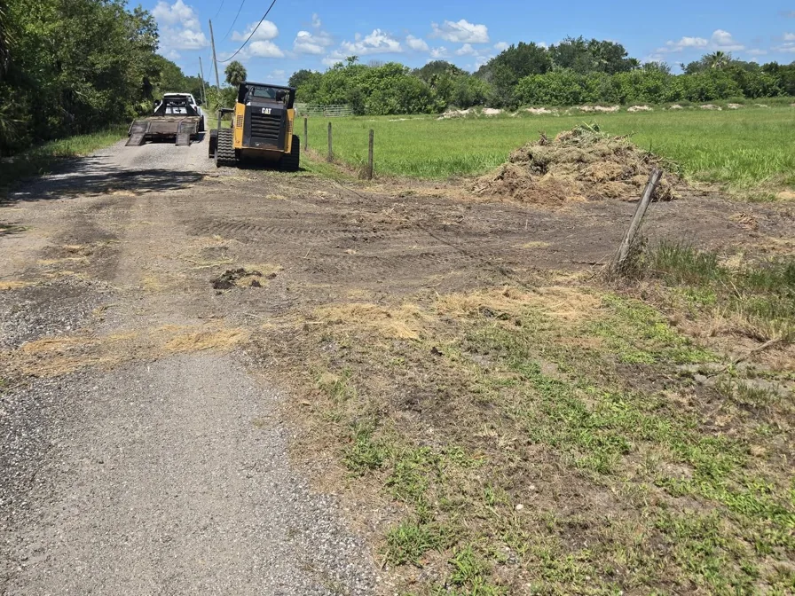 Cleared fence line beside a gravel road with the mulched growth piled and a track loader on the road, forestry mulching