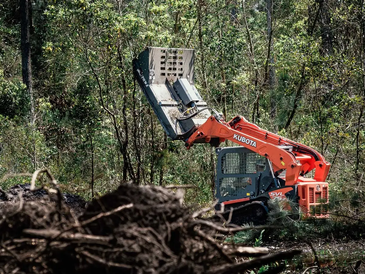 Track loader raising a brush cutter deck into small trees on a wooded lot, forestry mulching and mowing