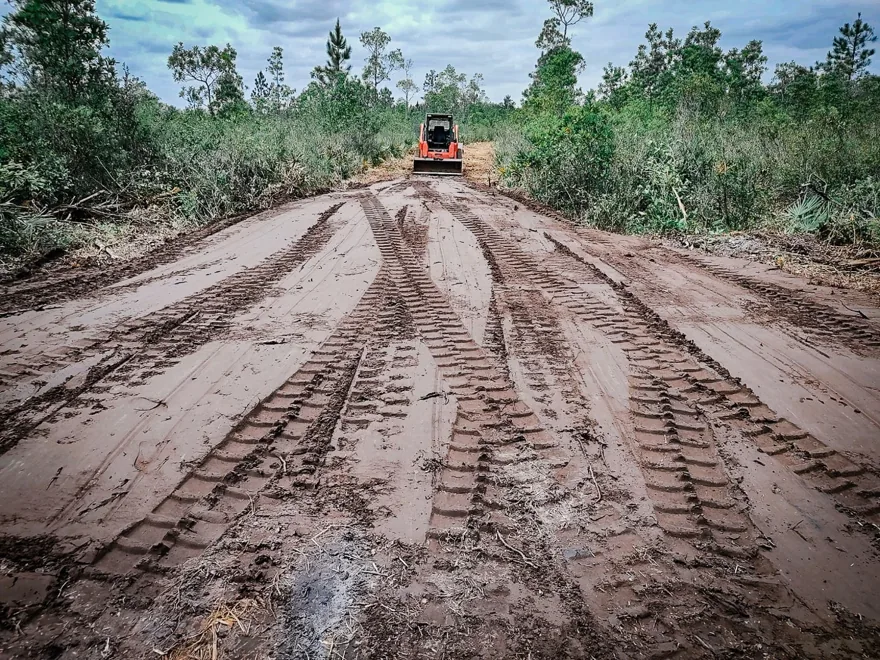 New dirt access road cut through palmetto scrub with a track loader at the far end, gravel driveways
