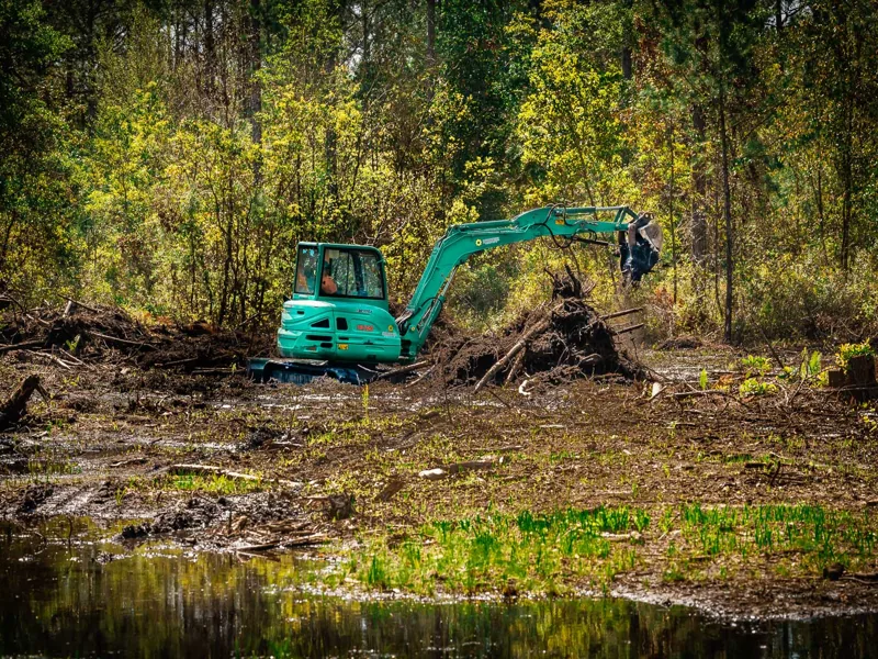 Excavator beside a pulled stump on a wet wooded lot, land clearing