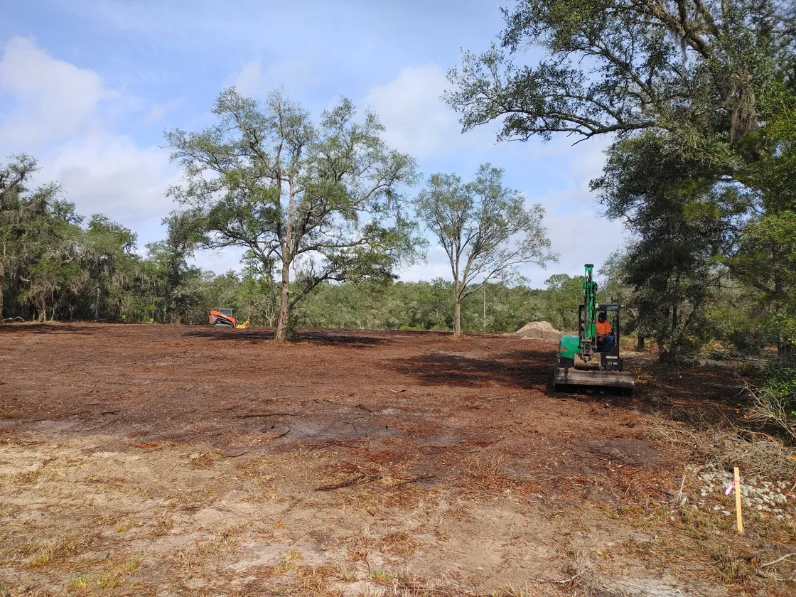 Mini excavator and a track loader working a cleared lot of bare dirt with live oaks left standing, land grading