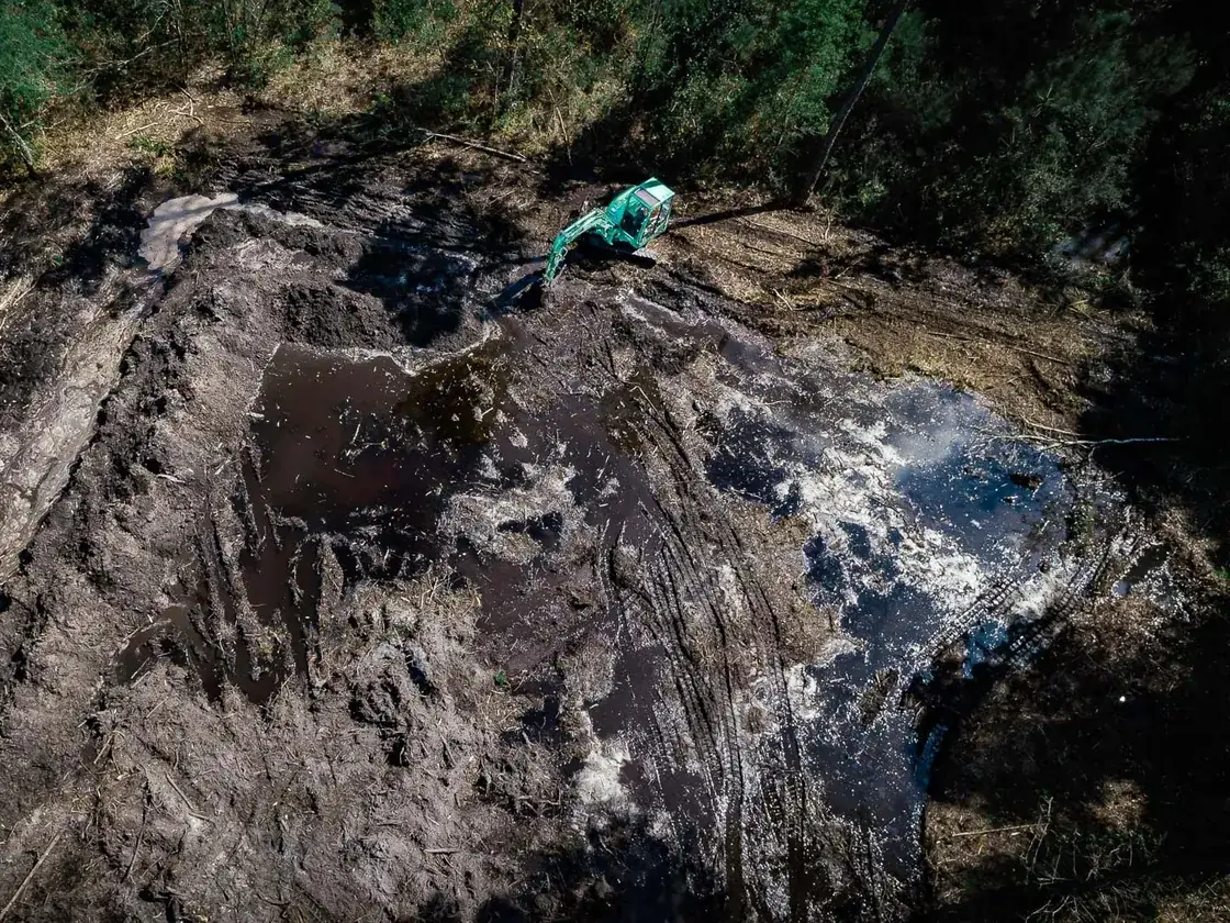 Overhead view of a new pond dig holding water while an excavator works the far end, pond construction