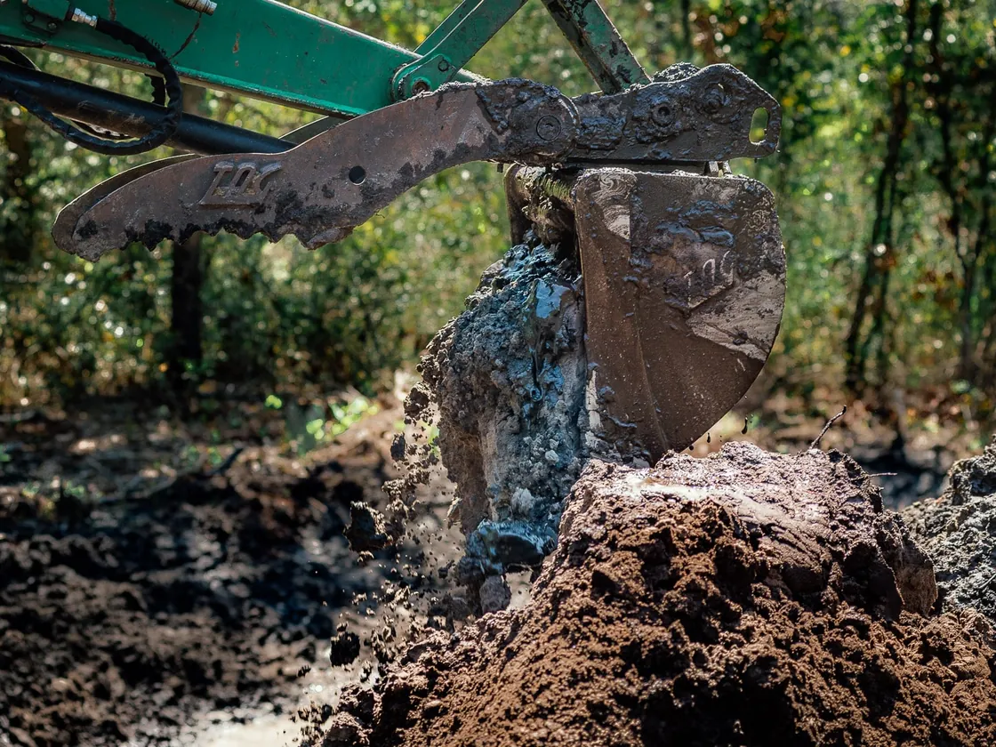 Excavator bucket and thumb lifting a muddy root ball out of a wet pond dig, pond construction