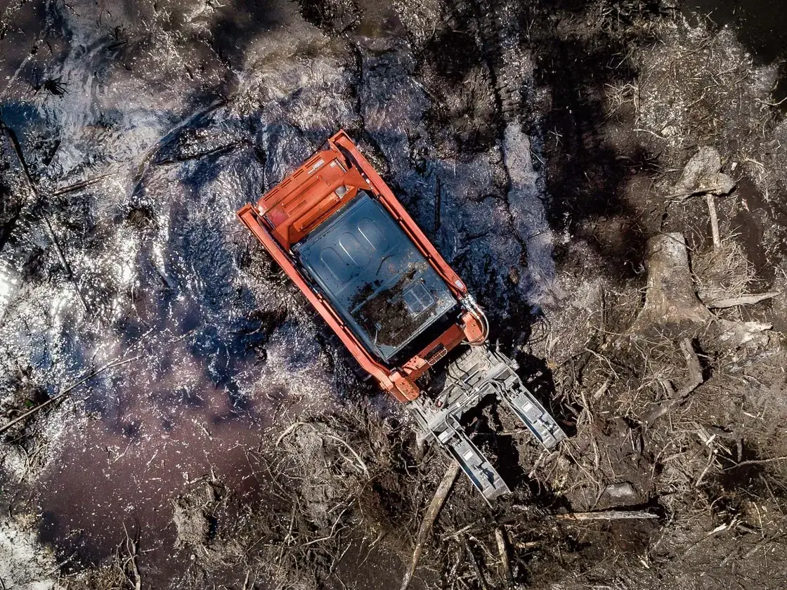Overhead view of a track loader with a tree puller clearing roots in a flooded pond dig, pond construction