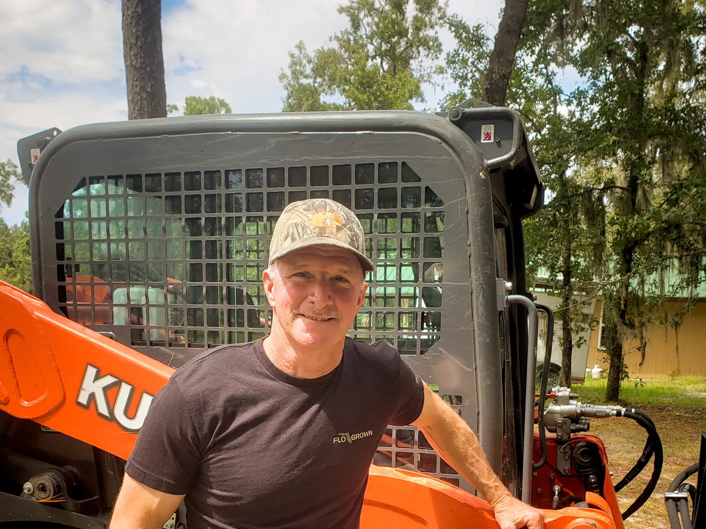 Smiling man in a camo cap beside a track loader, land clearing